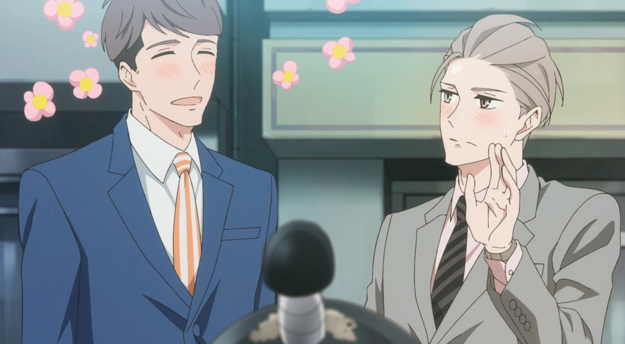 Momose, un hombre alto y moreno con un traje azul, se sonroja y se entusiasma con Shirosaki, un hombre rubio más bajo con una expresión inexpresiva y un traje color canela.